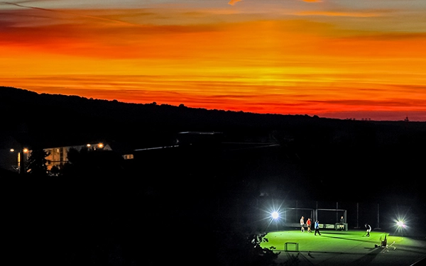 Hockeyspieler trainieren auf einem beleuchteten Spielfeld in der Dämmerung. Im Hintergrund leuchtet ein intensiver Sonnenuntergang in Orange- und Rottönen – Sportfoto von Michael Schalansky.